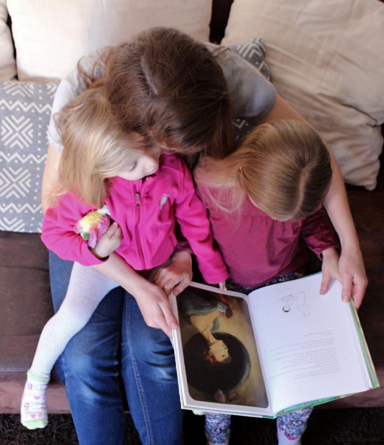 Mutter schaut mit ihren beiden Töchtern das Buch Mama von arsEdition an, zu sehen ist eine Illustration einer Frau mit großem Hut